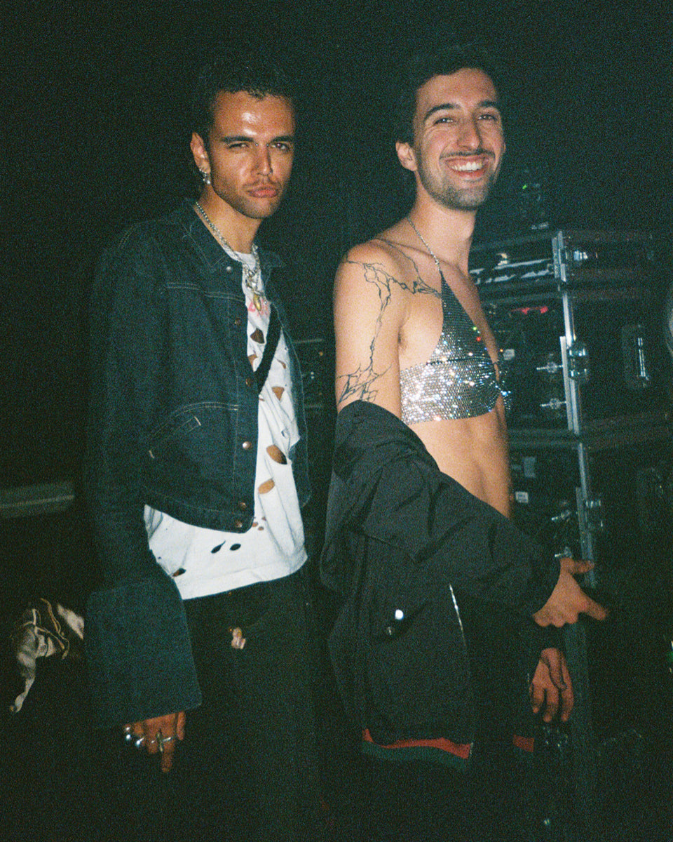 Two men standing together, one wearing a denim jacket and the other in a silver sequin top backstage at djg2g performance at roskilde festival