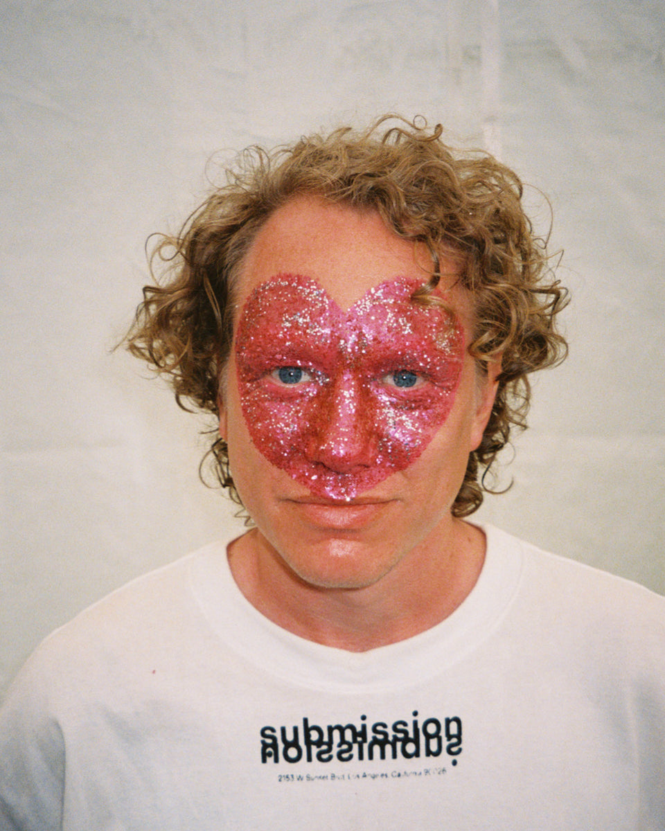 Person with a pink glitter heart on their face wearing a white shirt with submission beauty logo