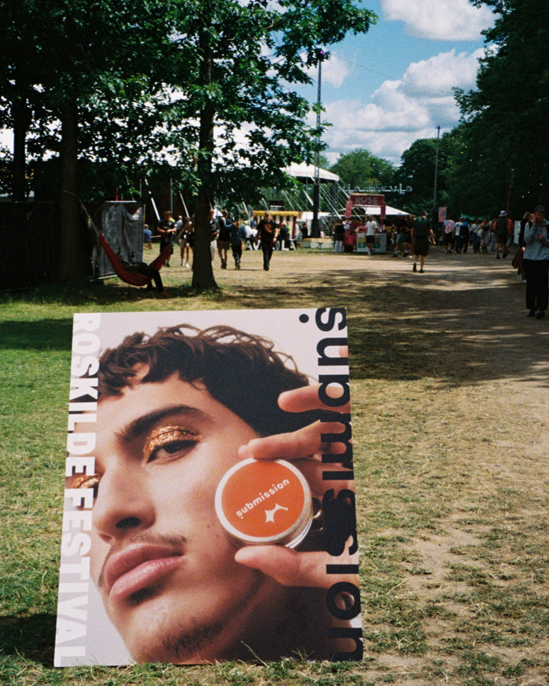 Poster of a person with Submission Beauty x Roskilde Festival limited edition glitter product on a grassy outdoor area