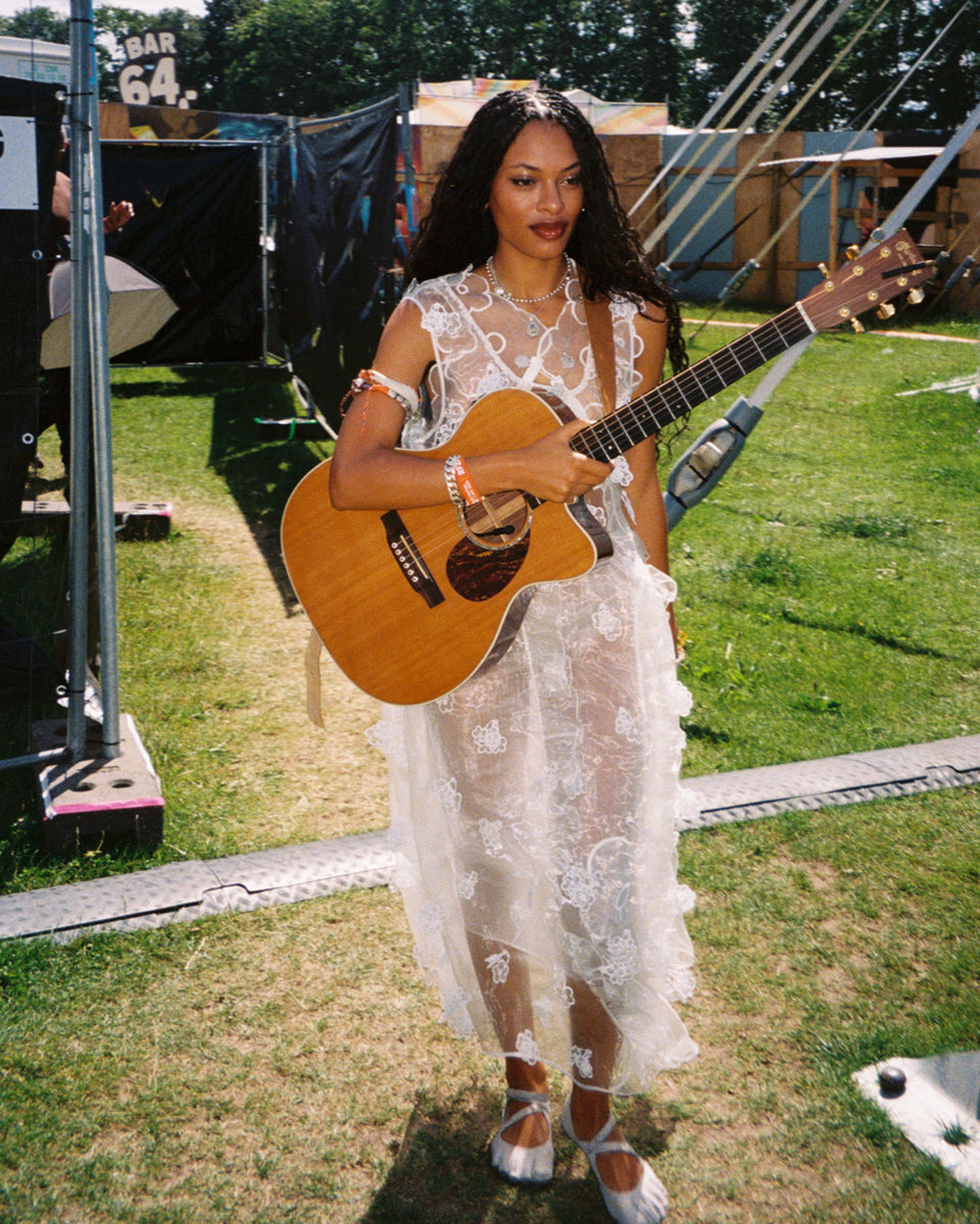 Artist annahstasia Enuke in a white lace dress playing an acoustic guitar outdoors.