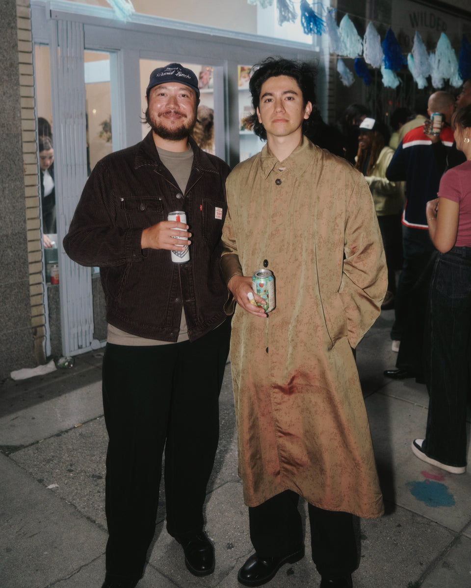Two men standing on a sidewalk, one in a dark jacket and cap, the other in a long beige coat, holding drinks.