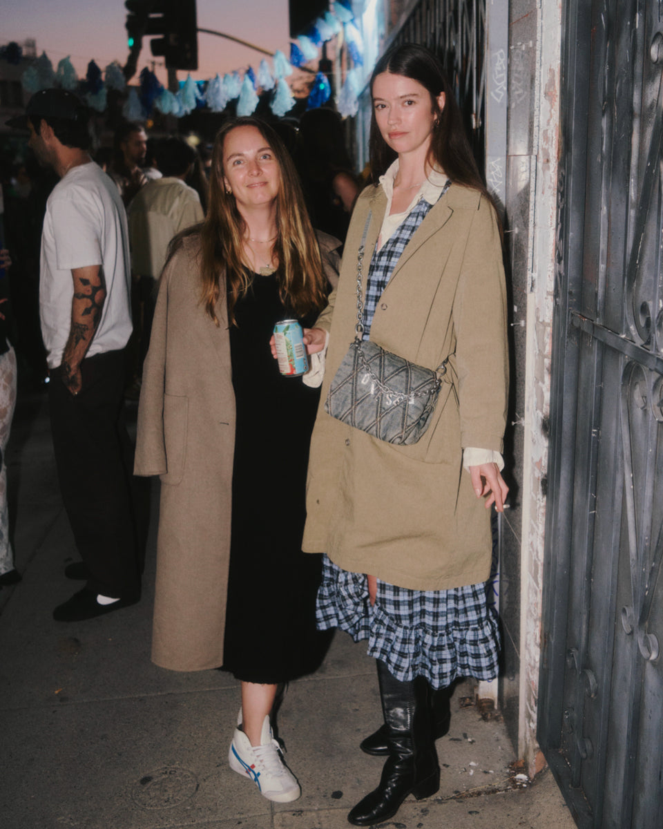 Two women standing together on a street, one holding a can, with a festive atmosphere in the background.