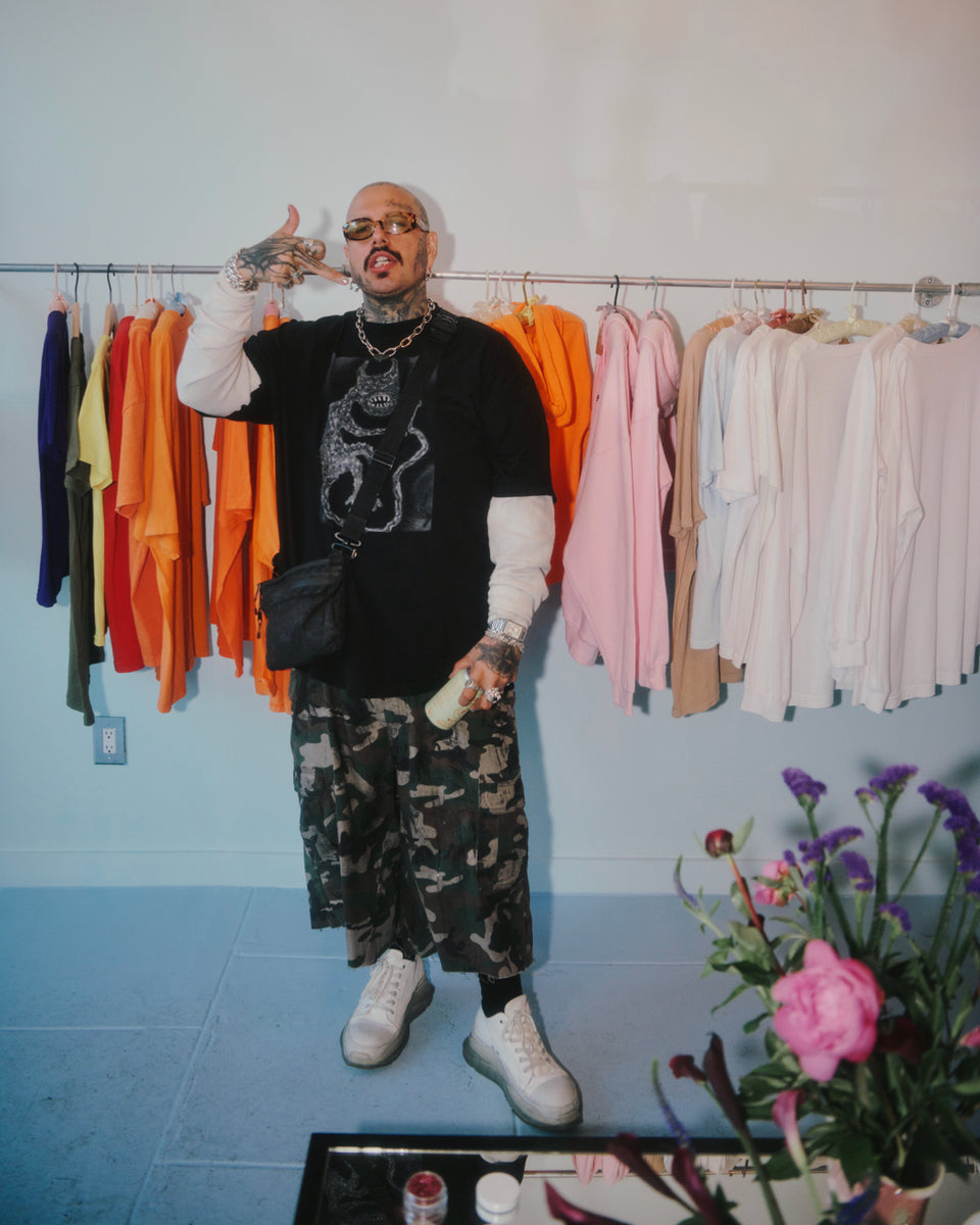 Person posing in a clothing store with racks of clothes and a vase of flowers on a table.