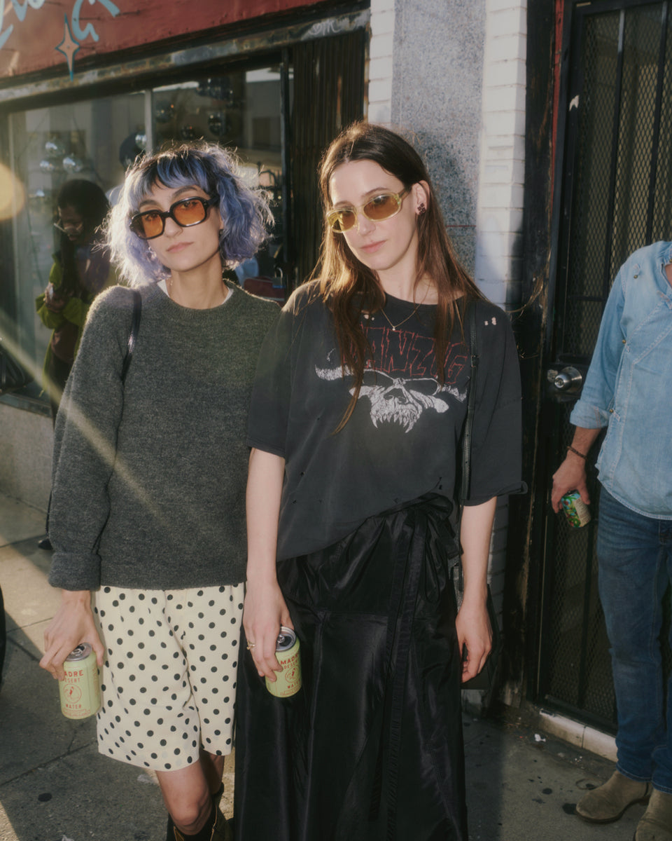 Two people standing on a street, one with purple hair and sunglasses, the other in a black outfit with a graphic design.