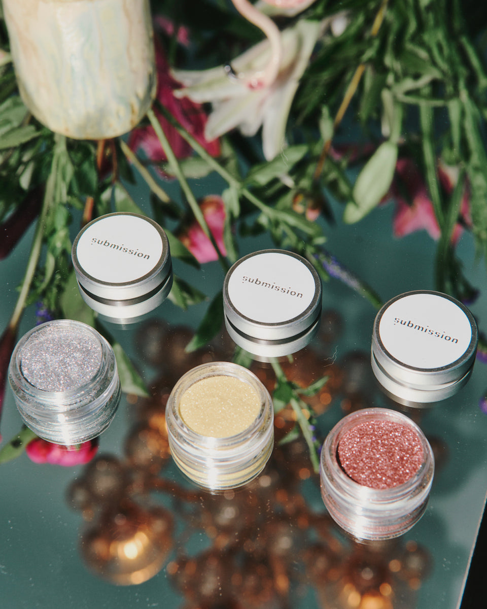 Set of makeup products with glittery finishes on a reflective surface with floral background