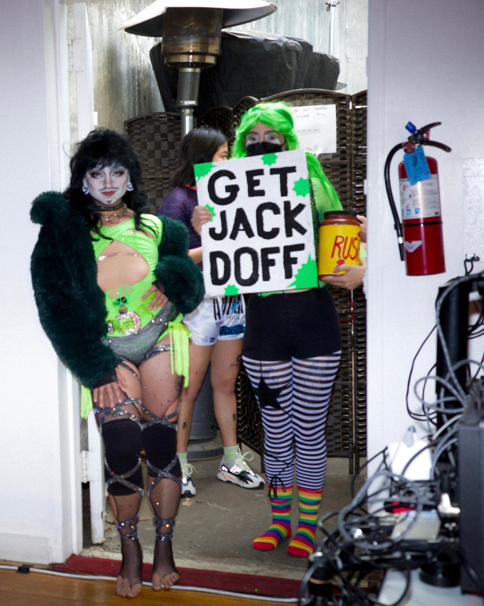 Person in green fur coat holding a sign with text, with camera equipment and fire extinguisher in the background.