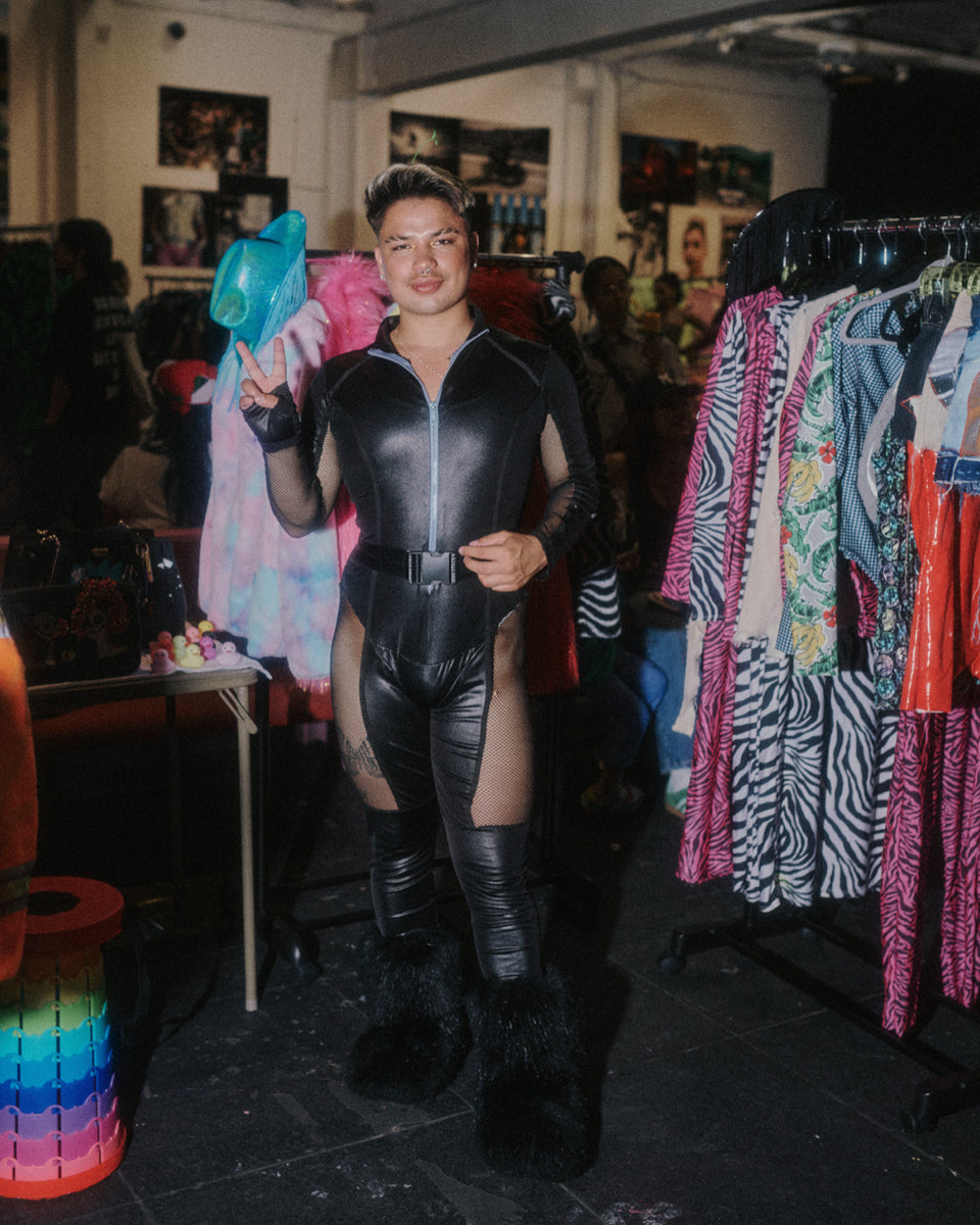 Person in a black leather outfit standing in a room with clothing racks and colorful items.