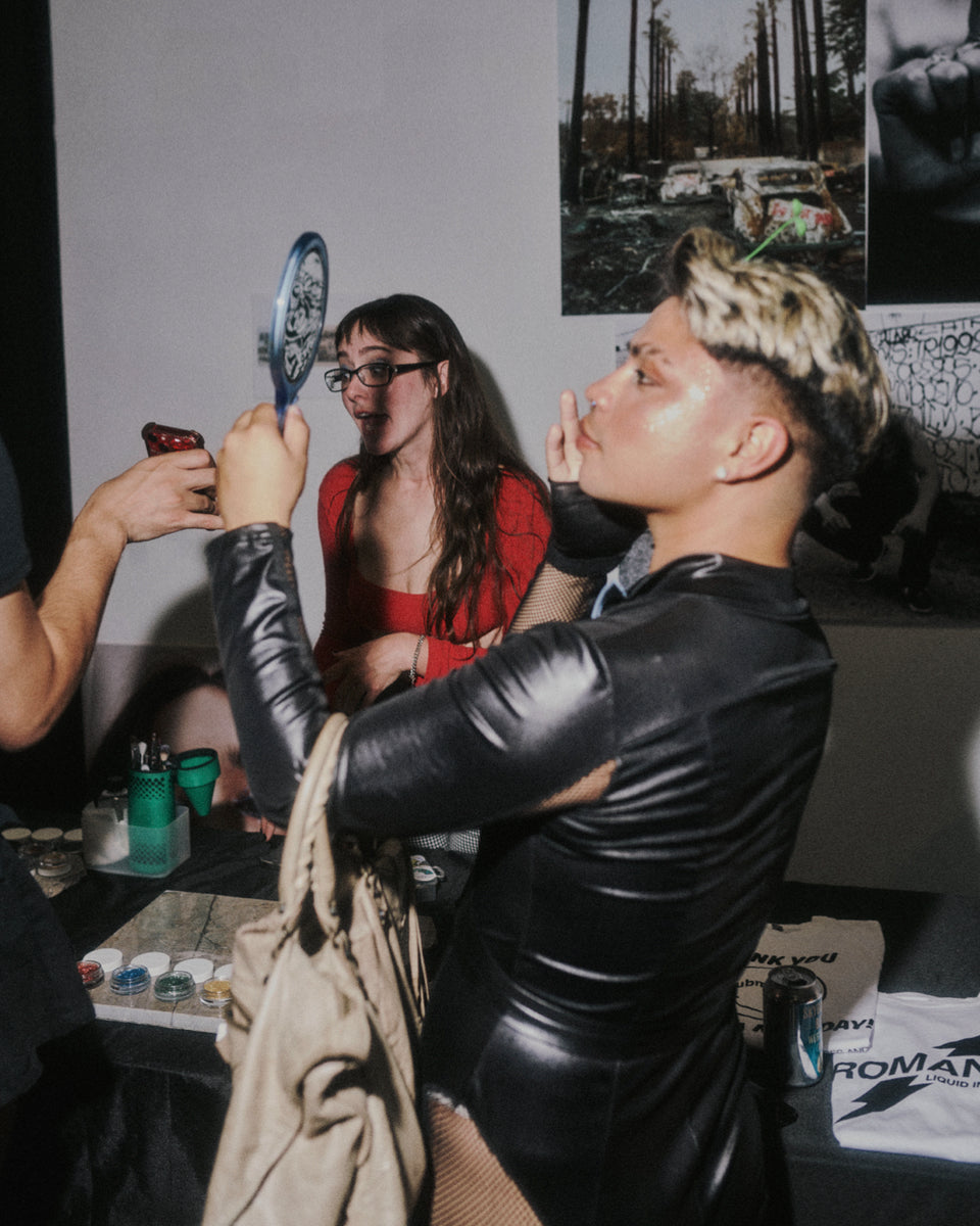 Person in a black leather outfit holding a paddle, with another person in a red dress in the background.