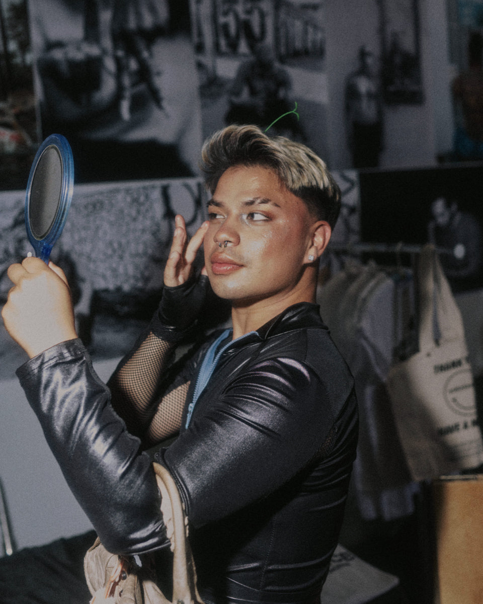 Person in a room with a mirror, wearing a black leather jacket and fishnet stockings.