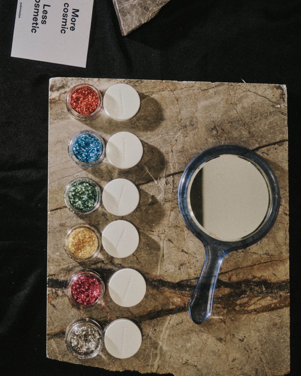 Set of Submission Beauty glitter products including a mirror and small containers on a textured surface.