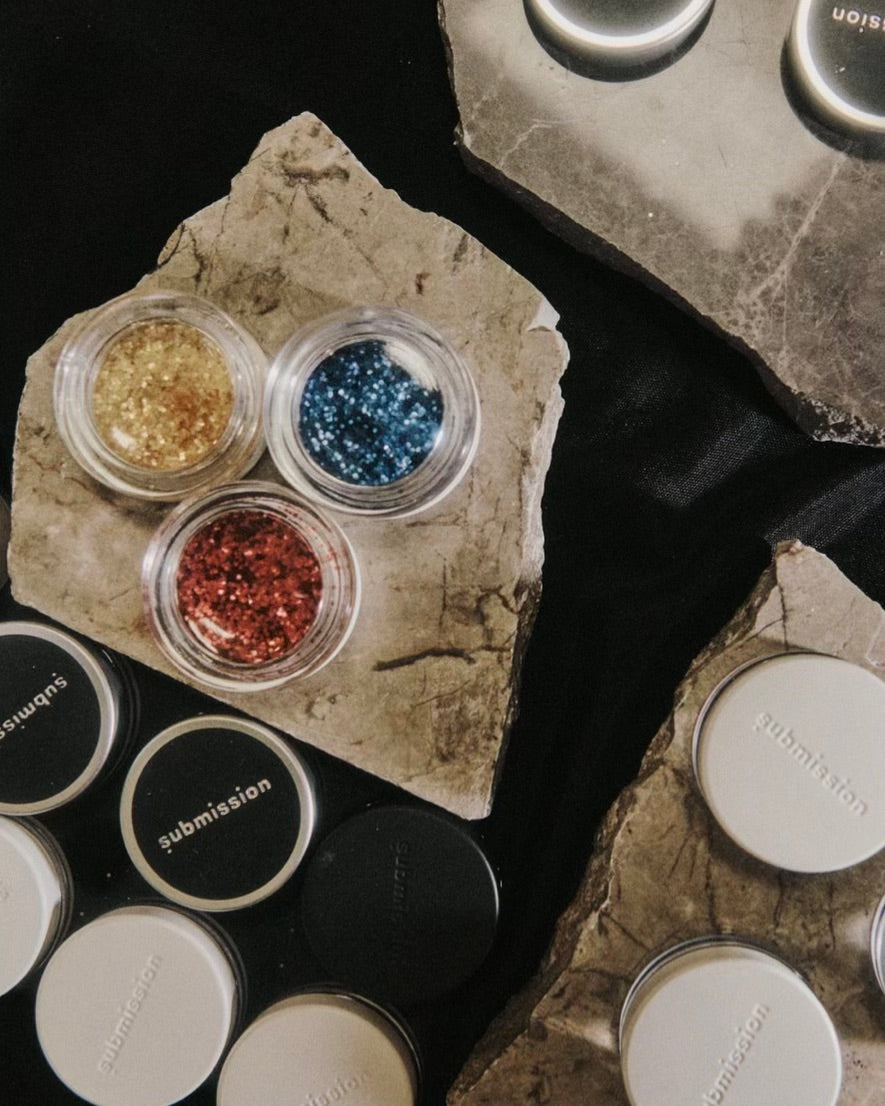 Three small containers of glitter on a stone surface with makeup products around.