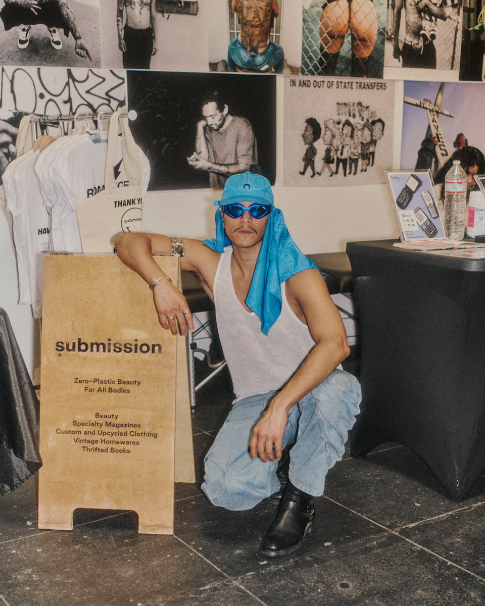 Person wearing a blue bandana and sunglasses sitting in front of a 'submission' sign and clothing rack.
