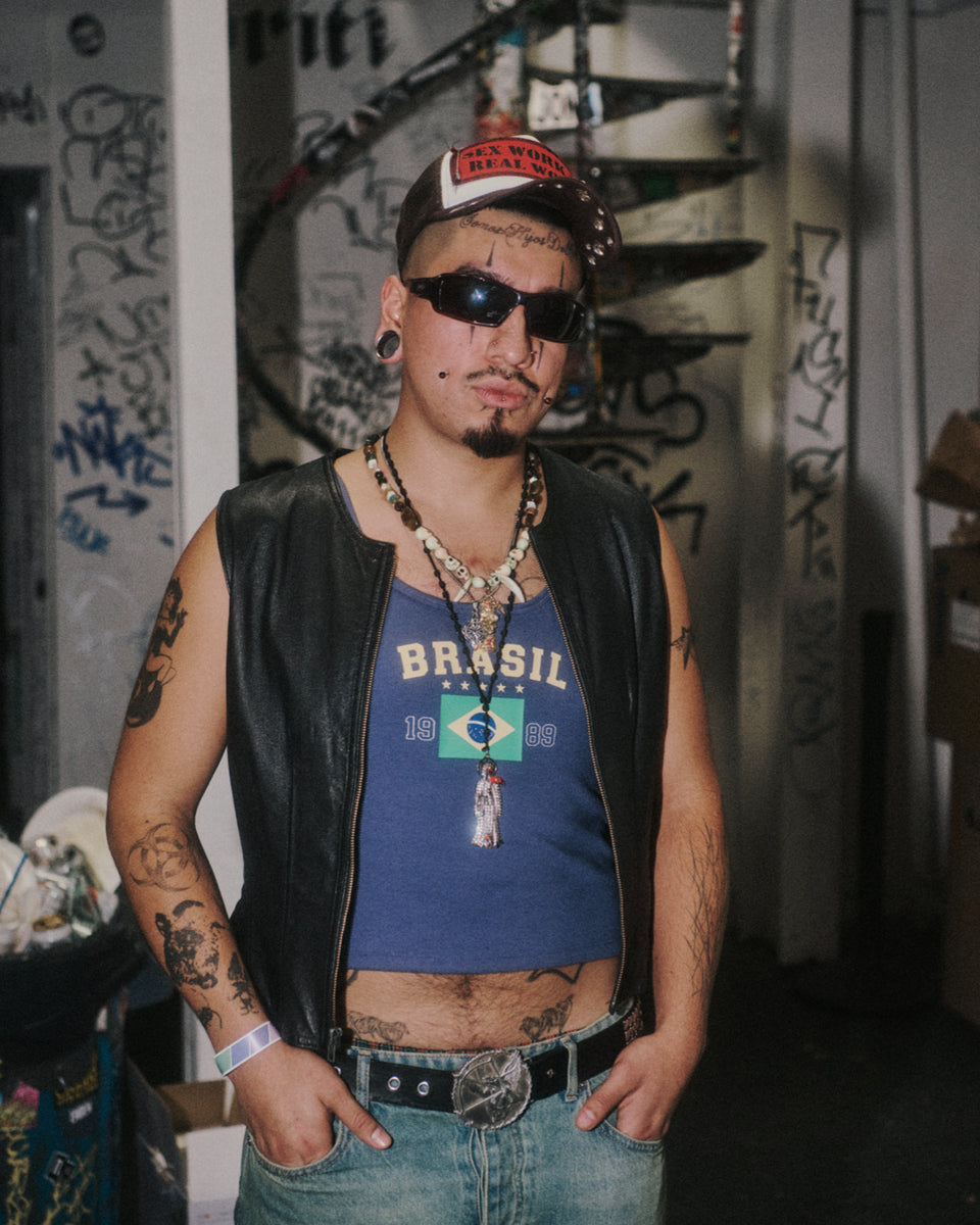 Man wearing a blue 'Brasil' t-shirt, black leather vest, and sunglasses in a graffiti-covered room.