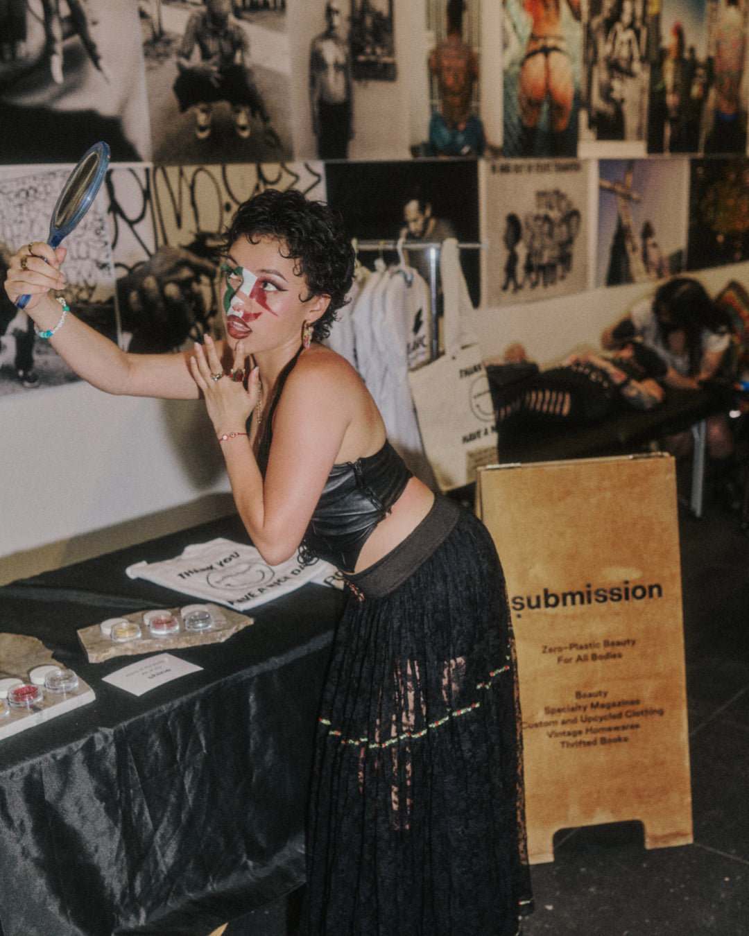 Woman applying makeup in front of a mirror with a 'submission' sign and people in the background.