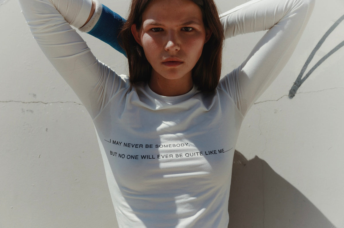 Person wearing a white long-sleeve shirt with text, standing against a light-colored wall.