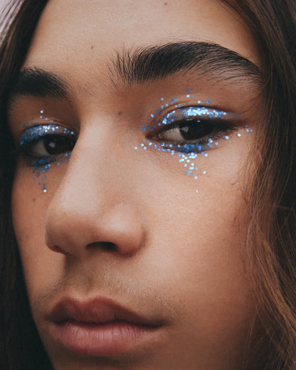 a cropped close-up of the face of a long-haired youthful male model with medium complexion and neutral expression, with flakes of plastic-free blue glitter around his eyes