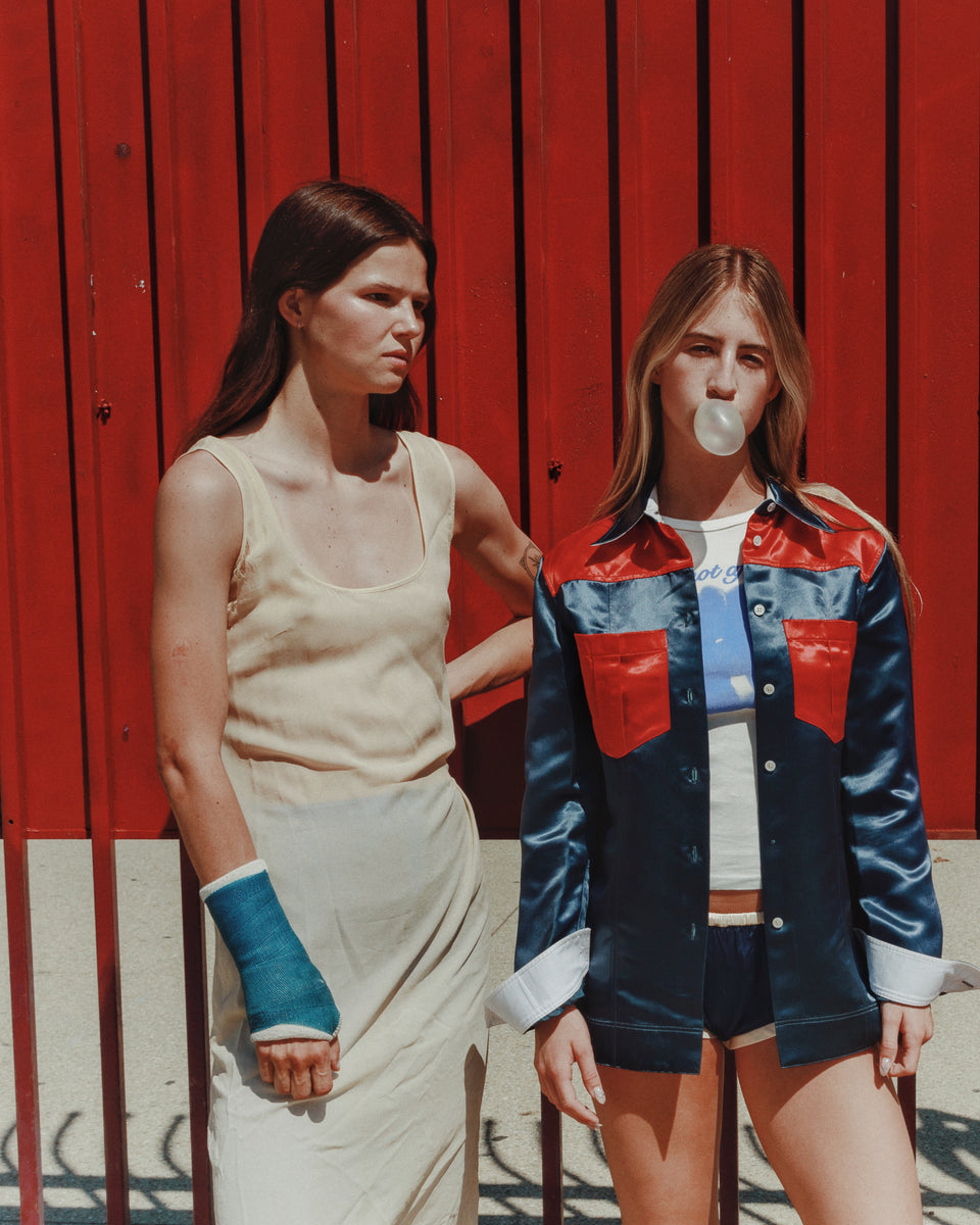 Two women standing against a red wall, one blowing a bubble with gum.