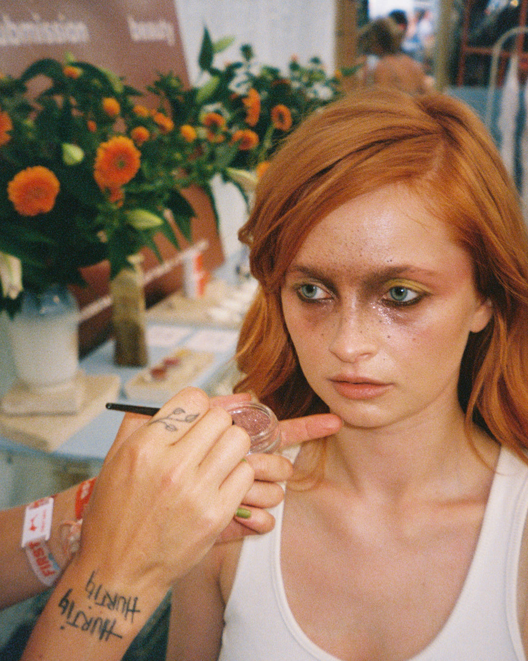 Person with red hair getting makeup applied at an event with floral decorations in the background