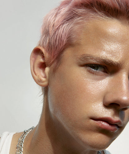 a cropped close-up of the face of a short-pink-haired youthful male model with light complexion and neutral expression, with Balm Shiny applied to his full face