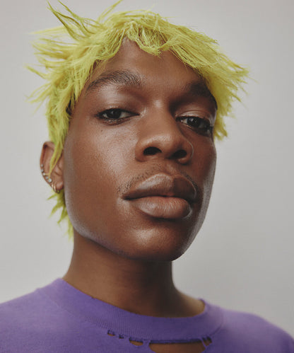 a cropped close-up of the face of a youthful male model  with medium-dark complexion, short dyed yellow hair and a purple t-shirt  with  a neutral expression, with Highlighter Deep applied to his eyes and lips and face