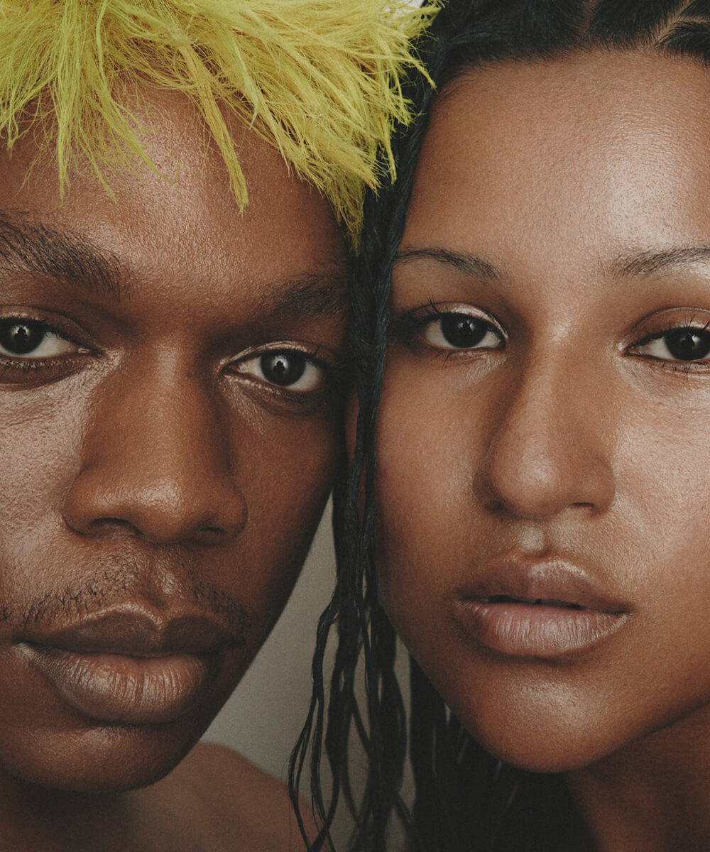 a cropped close-up of the face of  two youthful  models with medium-dark complexions cheek to cheek with  neutral expressions, with Highlighter Deep applied to their eyes and lips