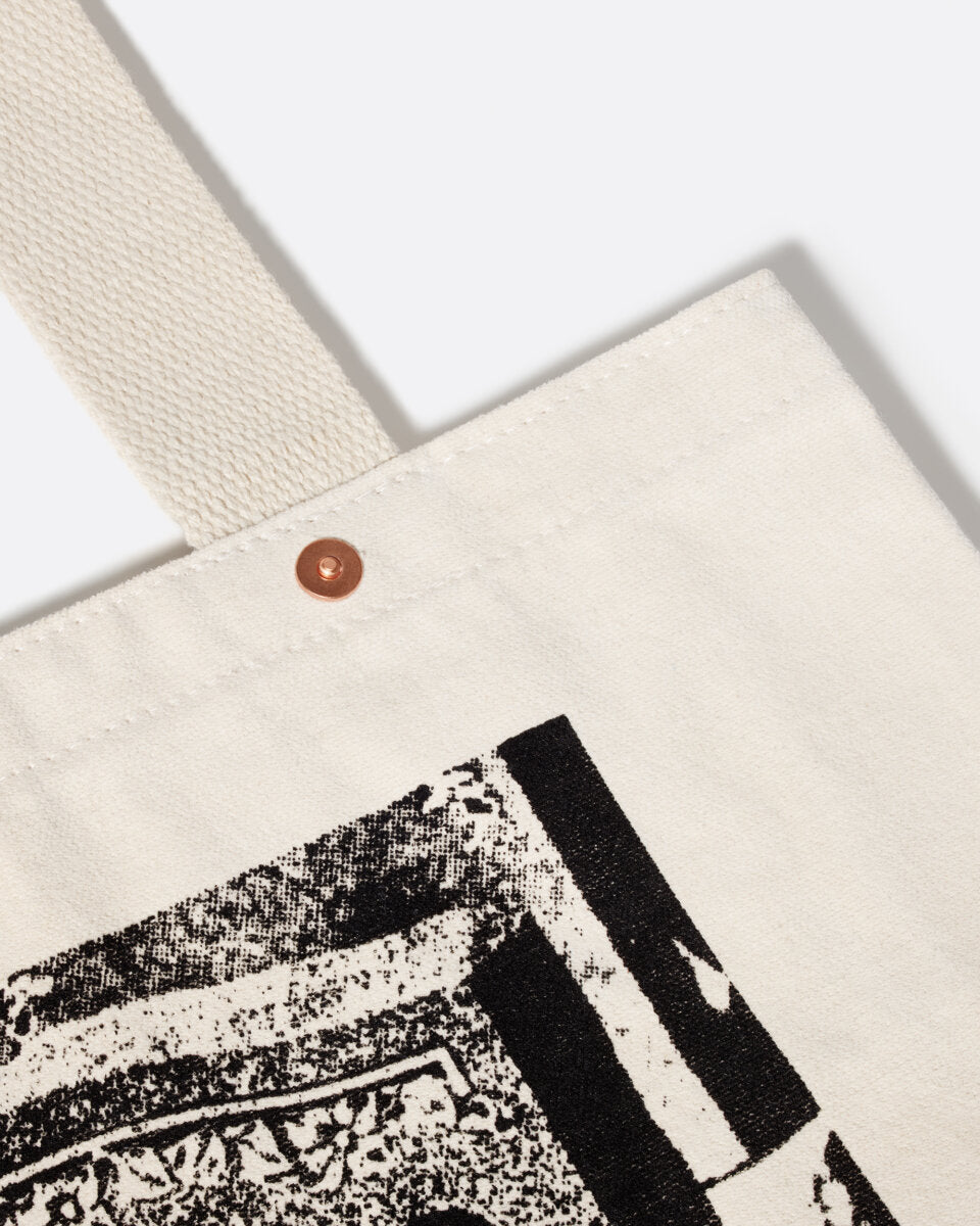 A cropped close-up product shot of the corner of an off-white tote bag with a large greyscale graphic in the center depicting a headstone with the words “PLASTIC IS OVER IF YOU WANT IT” in capital letters, and a decorative brass snap