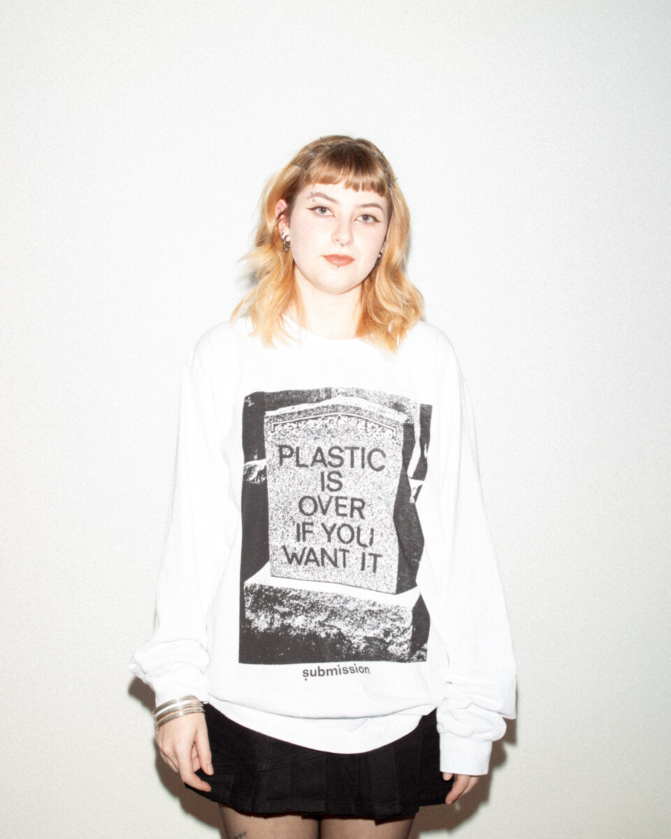 A portrait from the waist up of a young woman , wearing a white long-sleeved crewneck T-shirt with a large greyscale graphic in the center depicting a headstone with the words “PLASTIC IS OVER IF YOU WANT IT” in capital letters, and a logo beneath the graphic with the word “submission” in bold, lowercase type