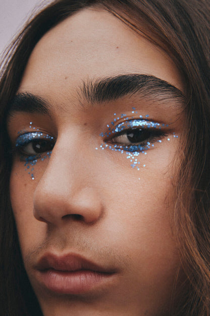 a cropped close-up of the face of a long-haired youthful male model with medium complexion and neutral expression, with flakes of plastic-free blue glitter around his eyes