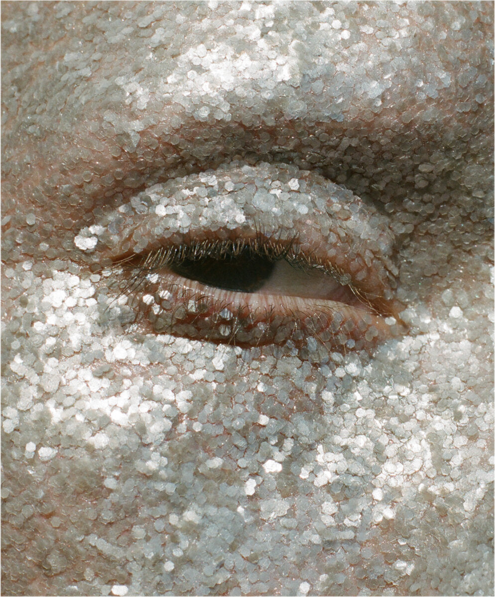 a cropped close-up of a partly open human eye, with flakes of plastic-free silver glitter coating all skin surrounding the eye including the upper and lower lids