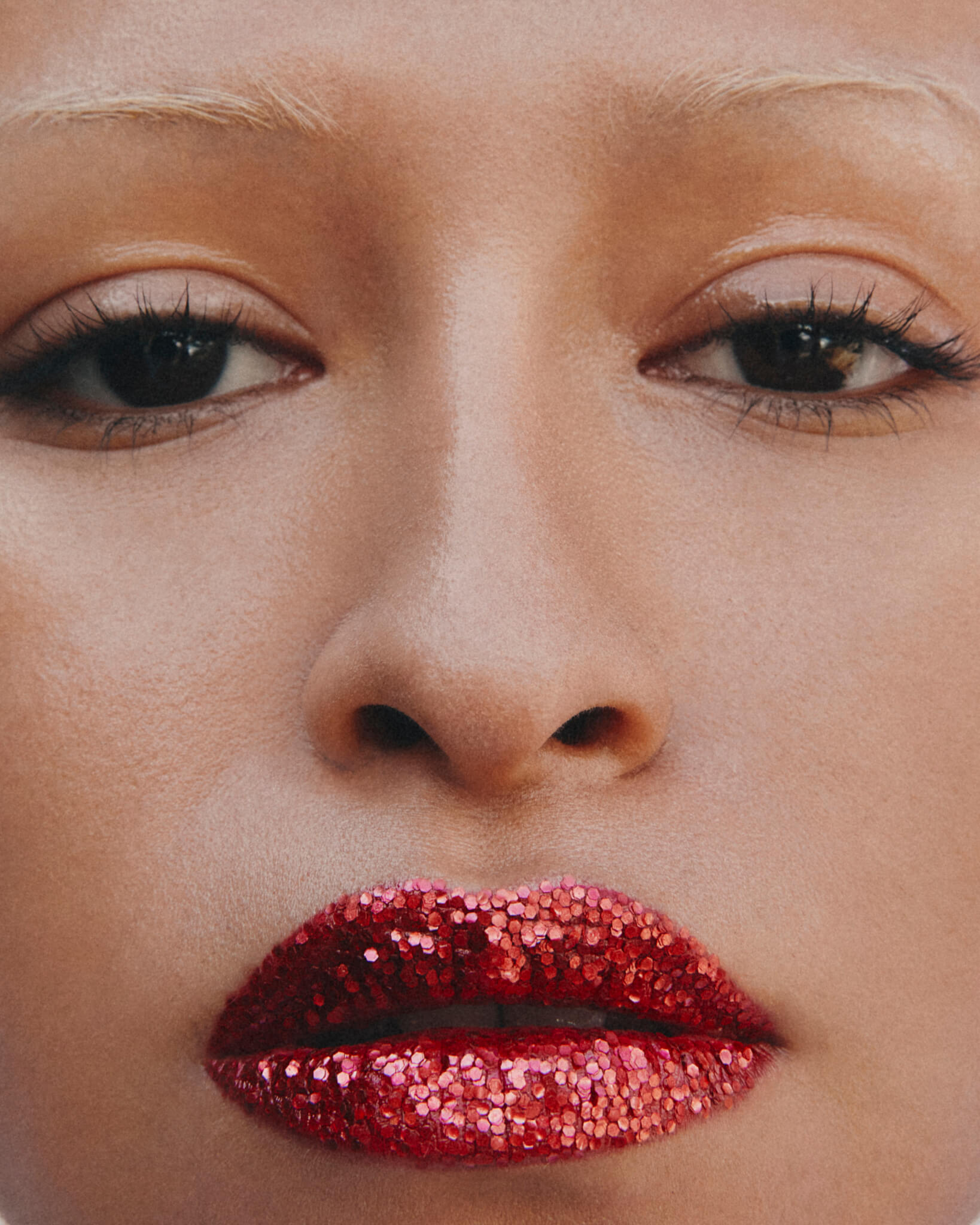 a cropped close-up of the face of a youthful female model with bleached eyebrows, medium complexion and neutral expression, with flakes of plastic-free red glitter coating her slightly parted lips