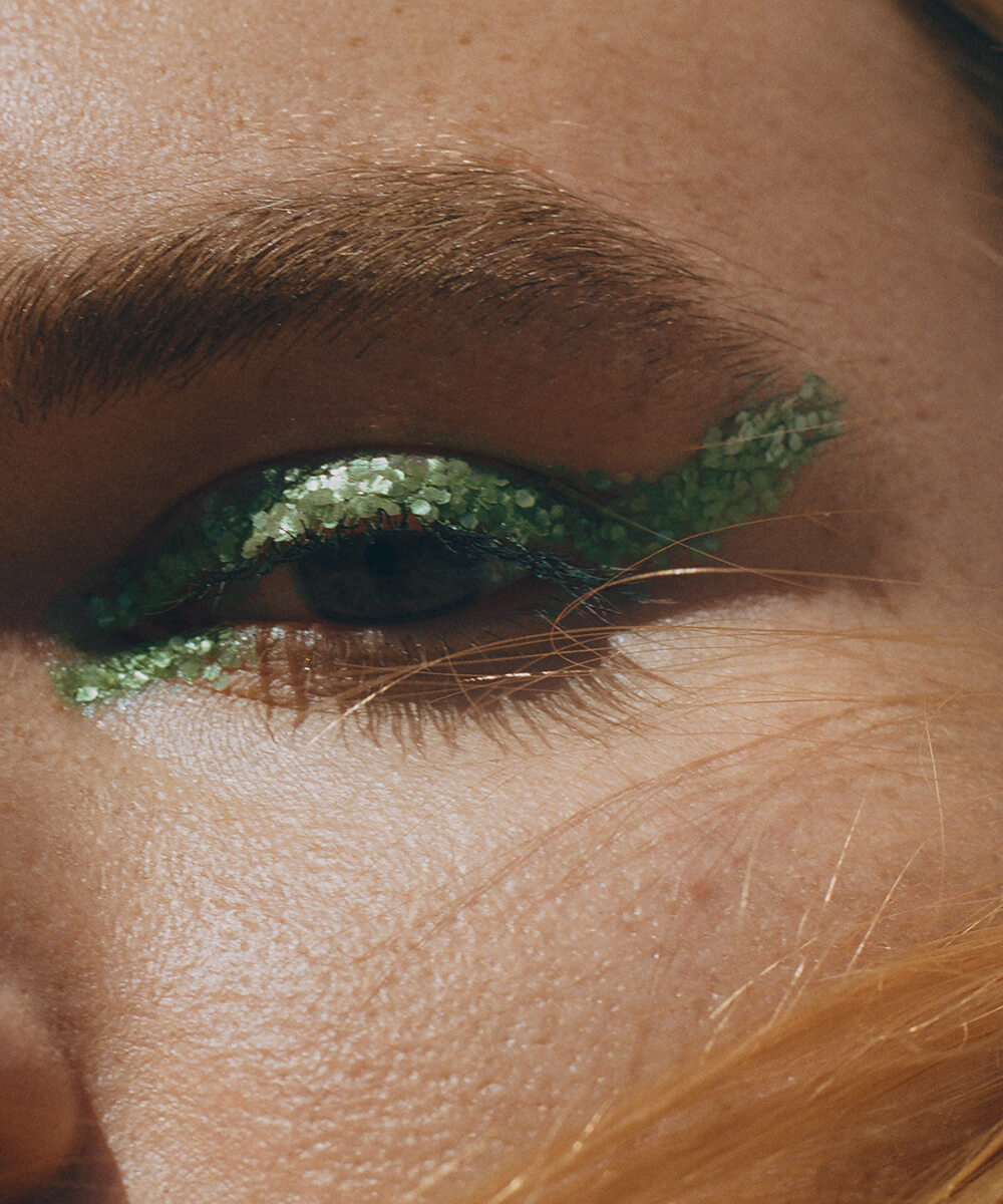 a cropped close-up of the left eye, brow, and cheek of a youthful model with medium complexion and a few dirty blonde hairs across her cheek, with flakes of plastic-free green glitter forming a cat-eye look across her upper eyelid and inner eye