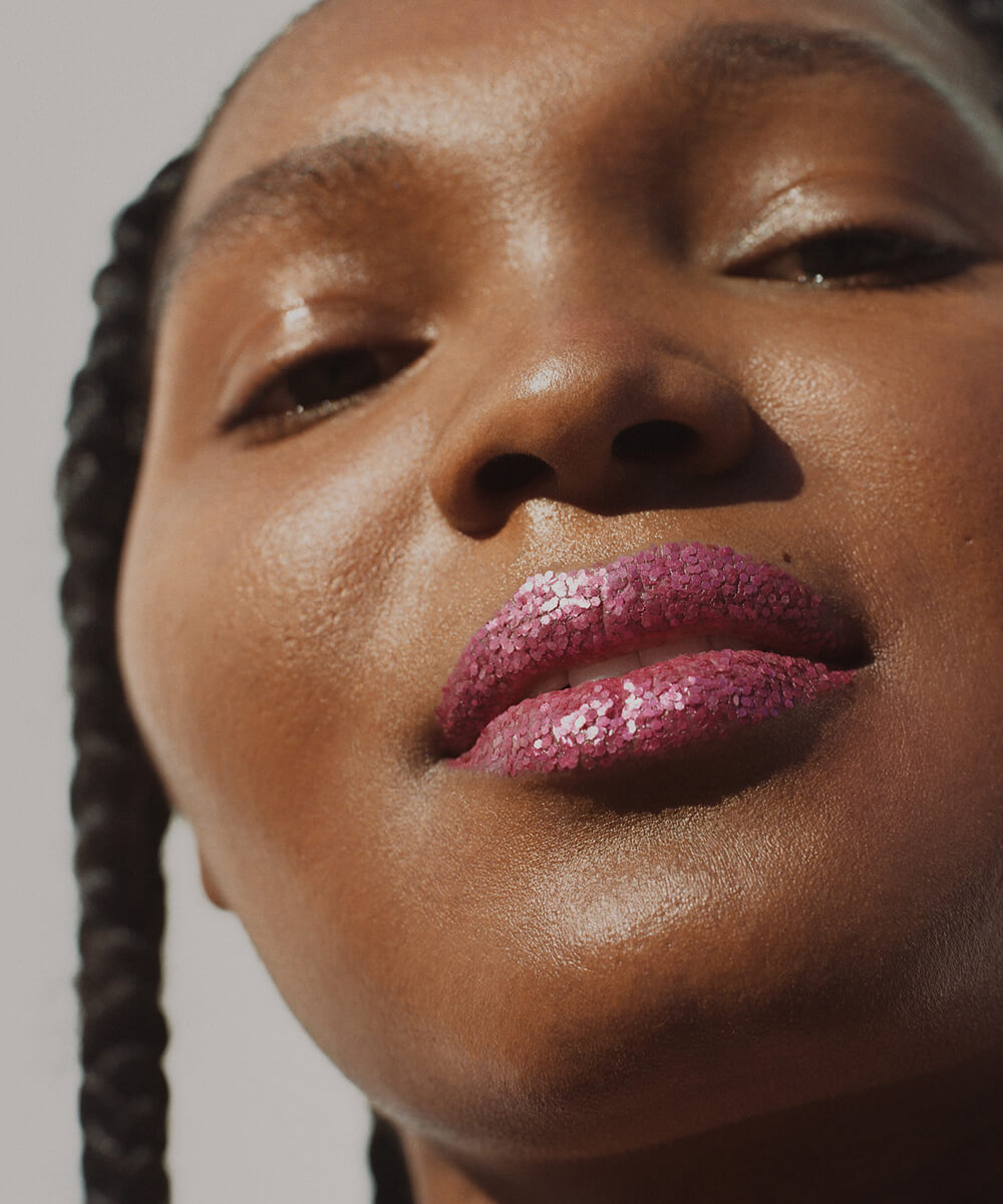 a cropped close-up of the face of a youthful female model with a deep complexion, raised chin and and slight smile, head cocked slightly to the right, and a single braid visible on her right side, with flakes of plastic-free pink glitter covering her lips