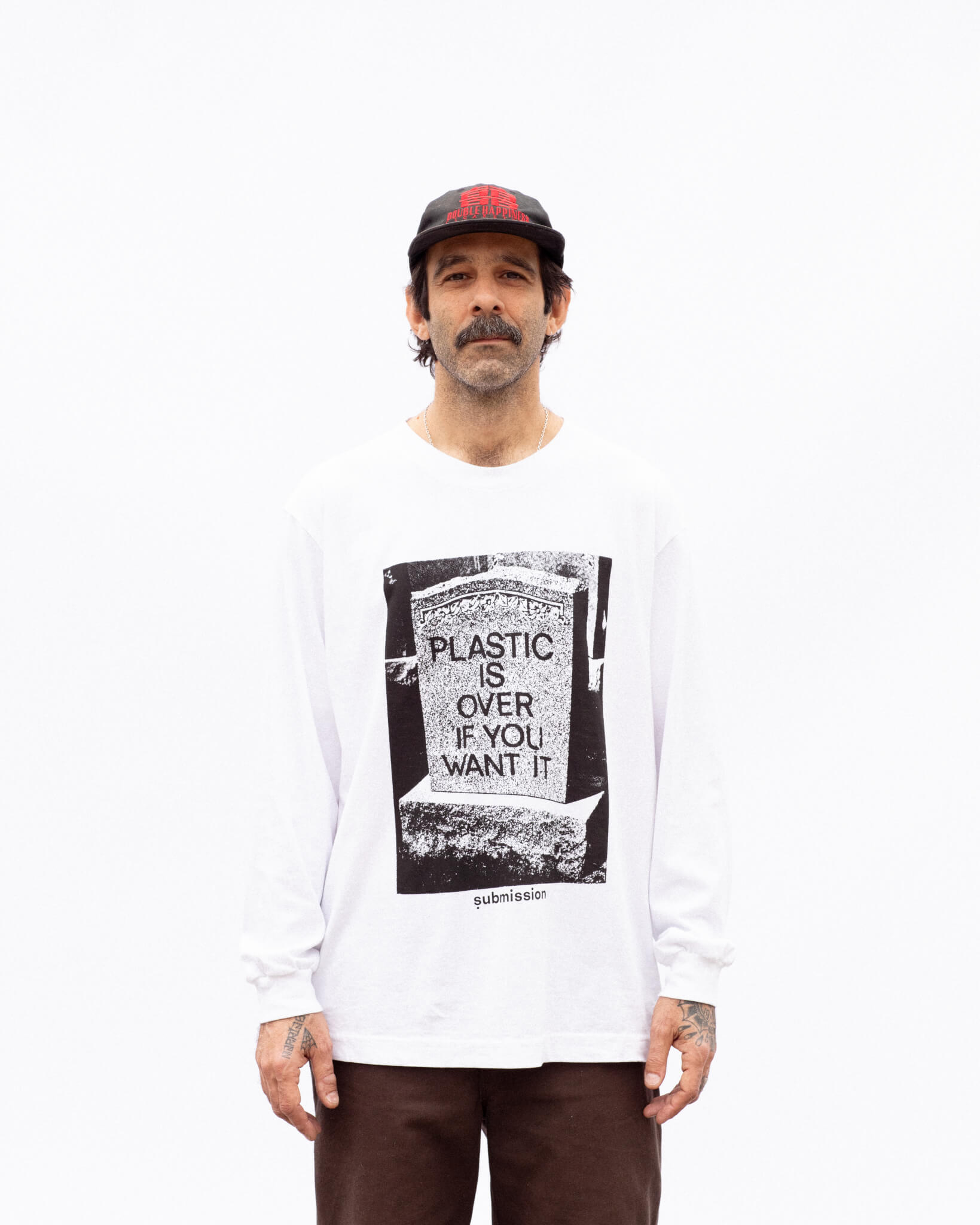 A portrait from the waist up of a middle-aged, male-presenting model with medium complexion standing in front of a white background, with a dark mustache and a black baseball cap with red branding, wearing a white long-sleeved crewneck T-shirt with a large greyscale graphic in the center depicting a headstone with the words “PLASTIC IS OVER IF YOU WANT IT” in capital letters, and a logo beneath the graphic with the word “submission” in bold, lowercase type