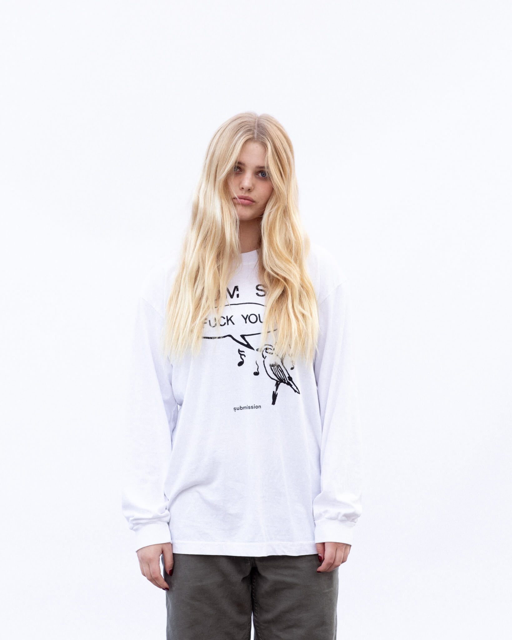 A portrait from the waist up of a young, female-presenting model with a pale complexion, long blonde hair partly obscuring her top, a white long-sleeved crewneck T-shirt with a black-and-white graphic in the center depicting a cartoon bird surrounded by musical notes with a speech bubble in which the words “FUCK YOU” appear in capital letters in quotation marks, above which the words “MOM SAYS” appear in capital letters, and a logo beneath the entire graphic with the word “submission” in bold, lowercase type