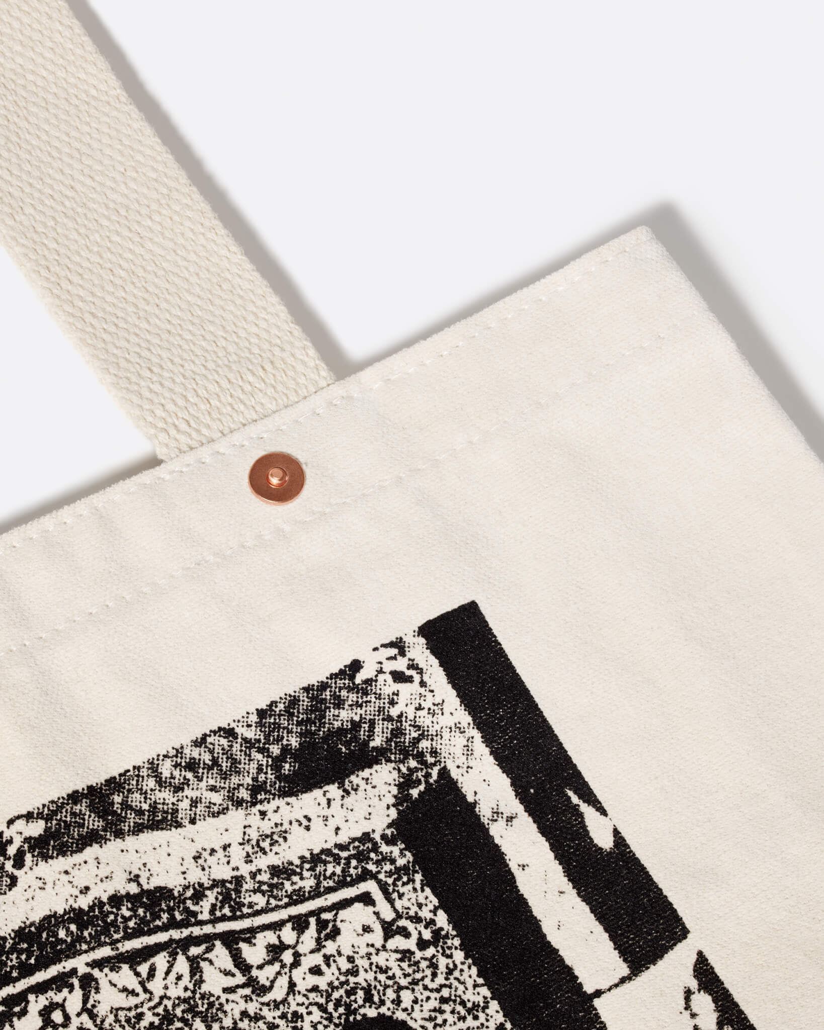 A detail shot of the top right corner of a brushed canvas tote bag made with 100% post-consumer recycled cotton, angled to the viewer’s left, showing the corner of a large greyscale graphic, and a coppertone metal rivet at the base of the handle.